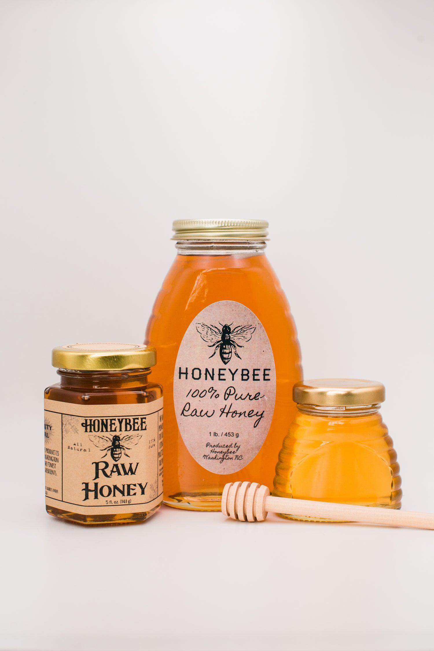 Three jars of premium honey are displayed against a white background. The jars vary in size and shape, with the labels reading "Honeybee Raw Honey." A wooden honey dipper lies in front of the jars, promising all-natural wildflower honey goodness inside.