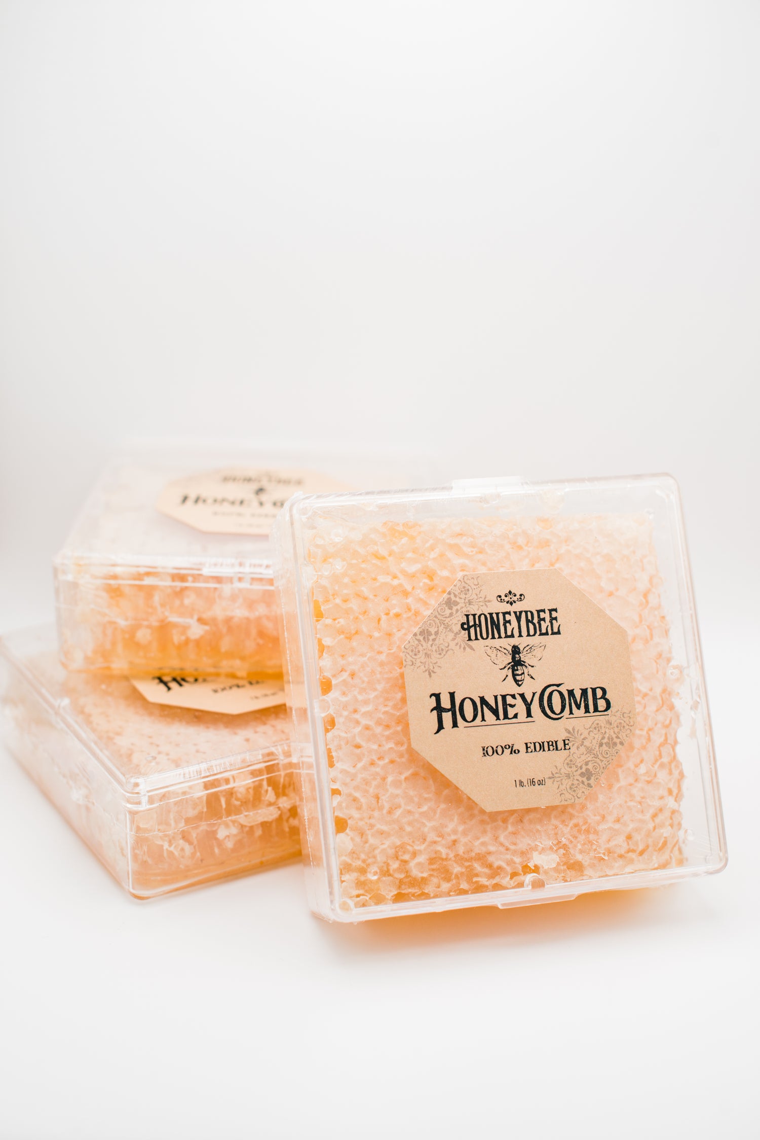 Three transparent plastic containers of pure raw honeycomb stacked on a white background. The label on the front container reads "Honeybee Honeycomb 1 lb. - 100% Edible" and "1 lb (16 oz)." The golden, textured honeycomb inside is perfect for charcuterie boards.