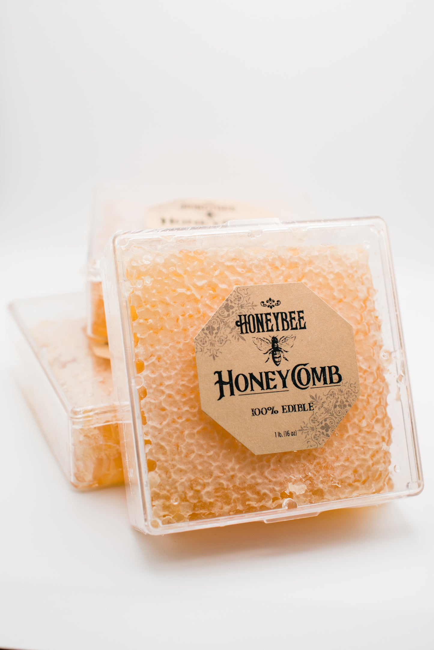 Three clear plastic containers filled with honeycomb are stacked. The front container shows a label that reads "Honeybee Honeycomb 1 lb." The golden, textured honeycomb inside each container is perfect for charcuterie boards or enjoying as pure raw honeycomb. The background is plain white.