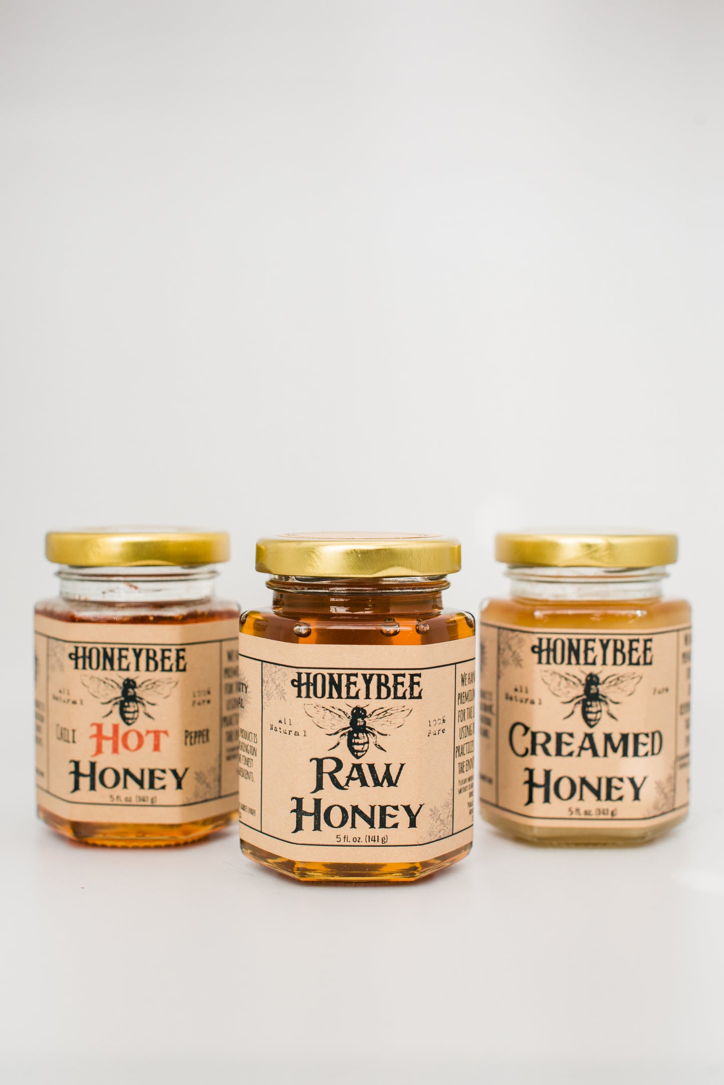 Three jars of Honeybee brand honey are displayed against a white background. From left to right, the labels read "Hot Honey," "Raw Wildflower Honey," and "Creamed Honey." The Hot Honey is infused with hot chili peppers. All jars have gold lids and beige labels with black and red text.