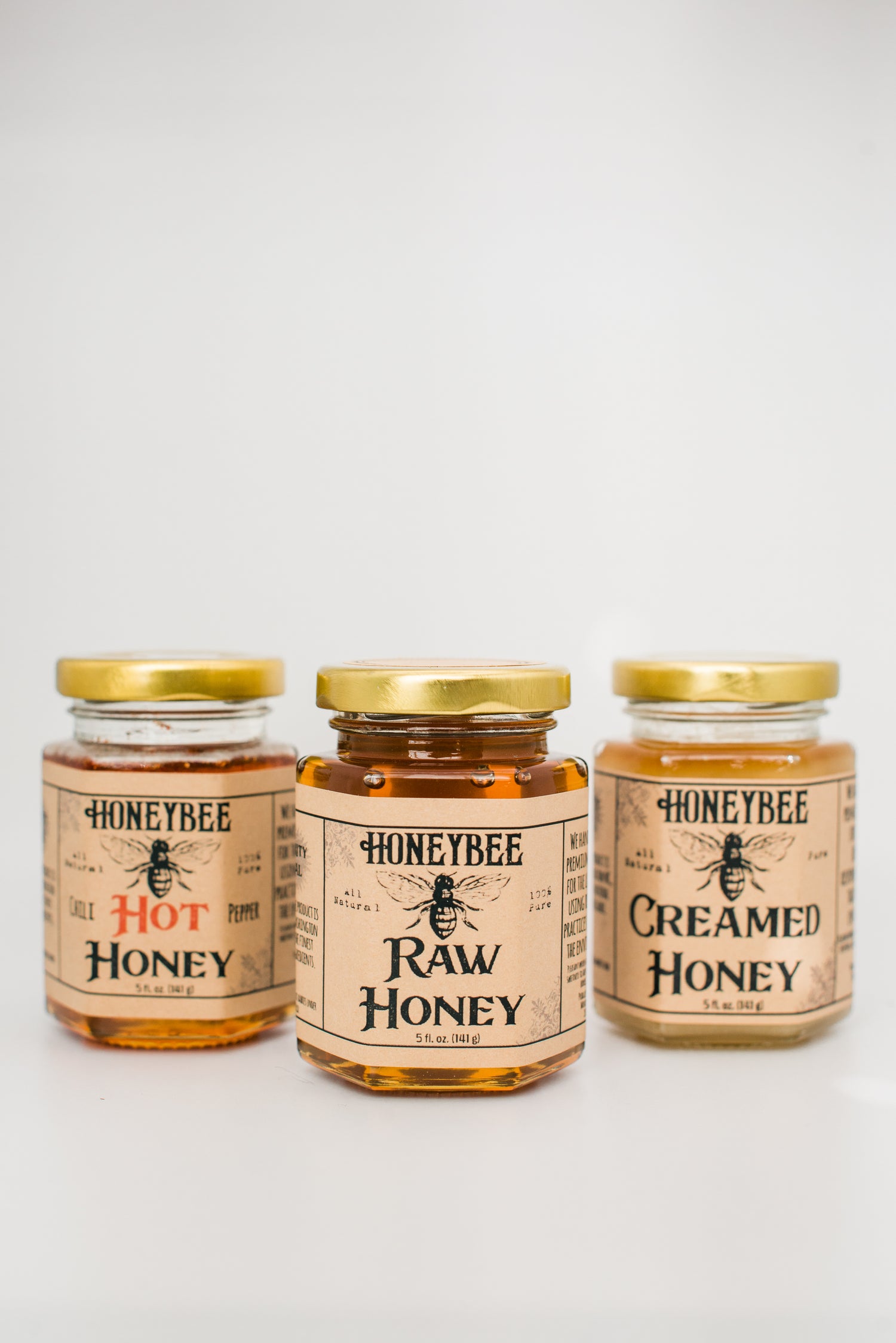 Three jars of Honeybee brand honey are displayed against a white background. From left to right, the labels read "Hot Honey," "Raw Wildflower Honey," and "Creamed Honey." The Hot Honey is infused with hot chili peppers. All jars have gold lids and beige labels with black and red text.